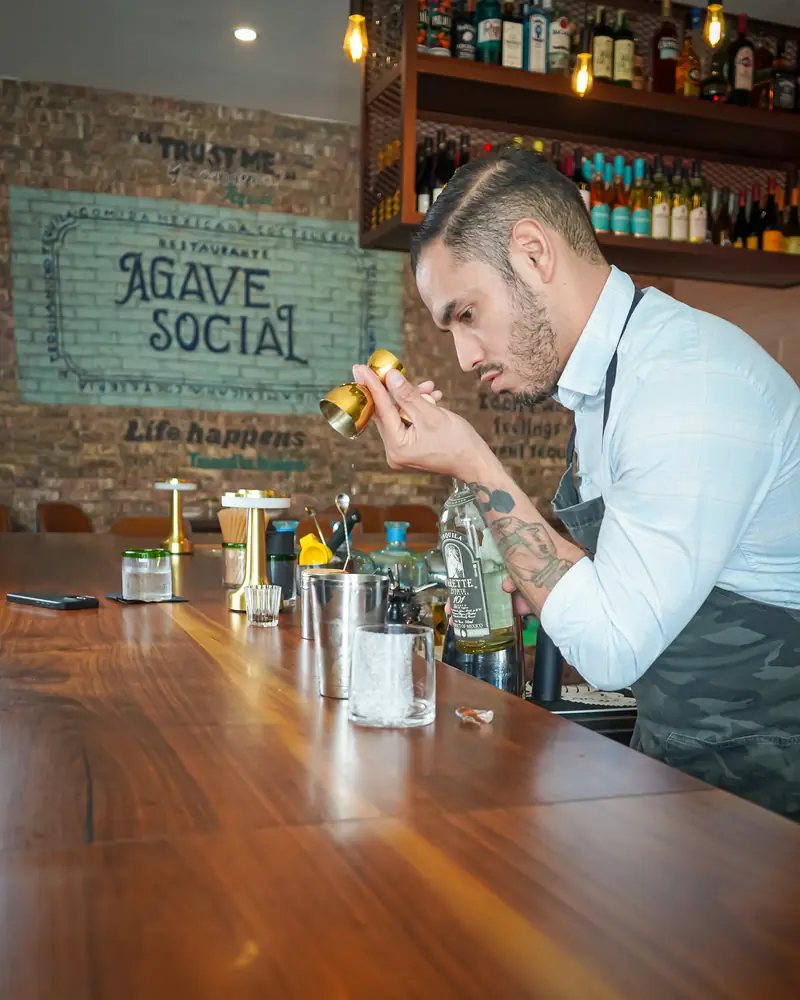 Bartender finishing a cocktail at the bar in front of the Agave Social brick mural