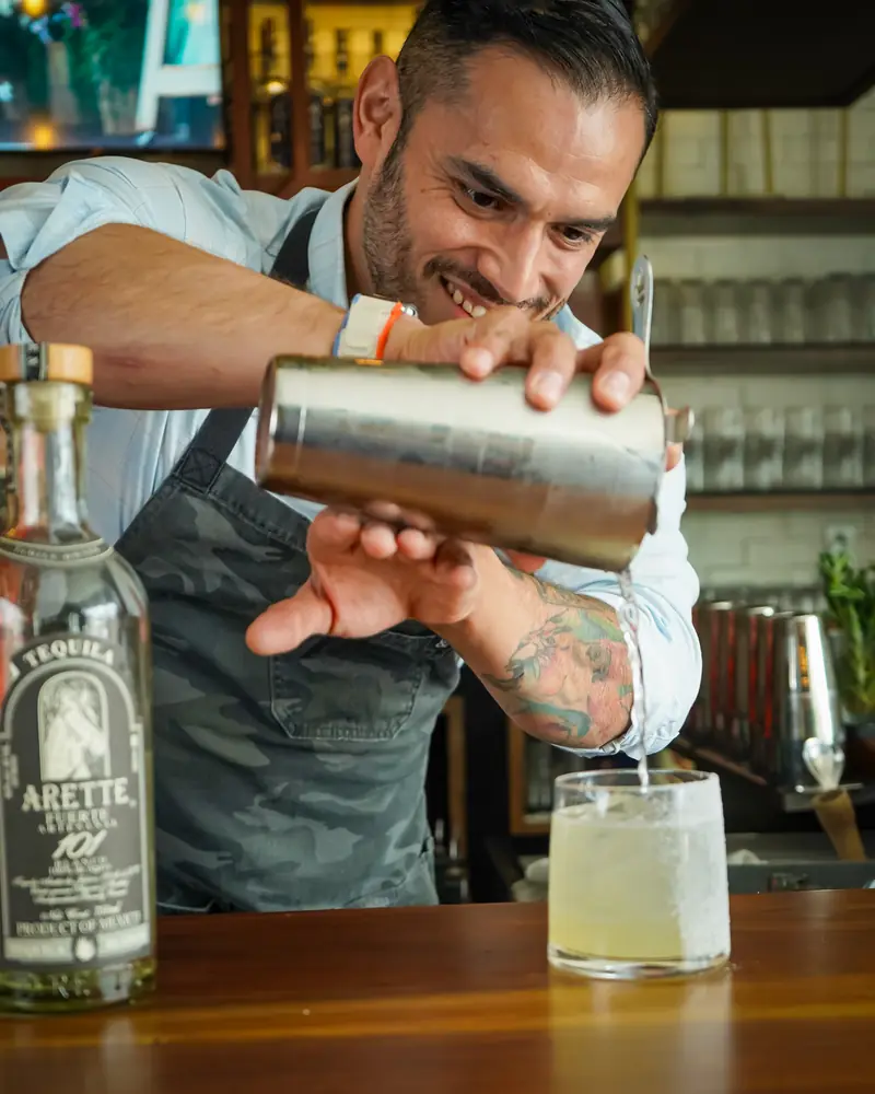 Bartender smiling as he pours from a cocktail shaker at the Agave Social bar, a bottle of Arette tequila beside him