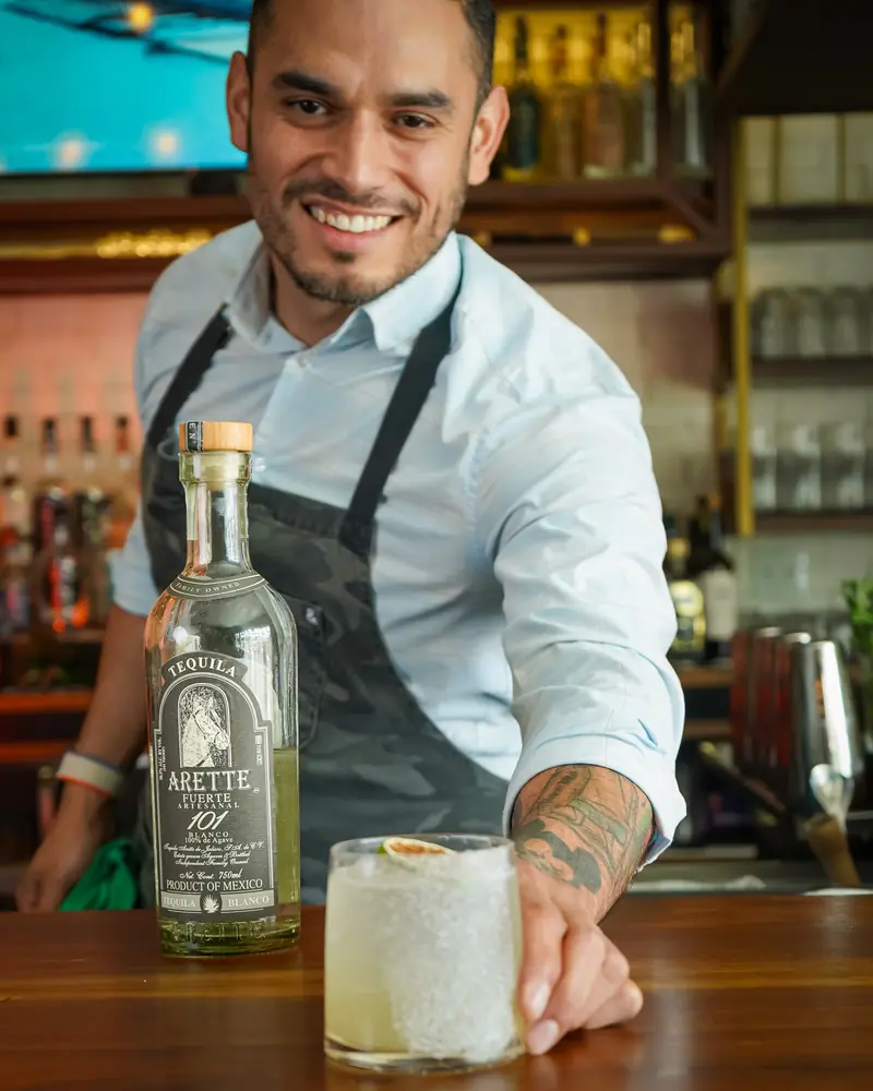 Bartender smiling as he slides a cocktail across the bar next to a bottle of Arette