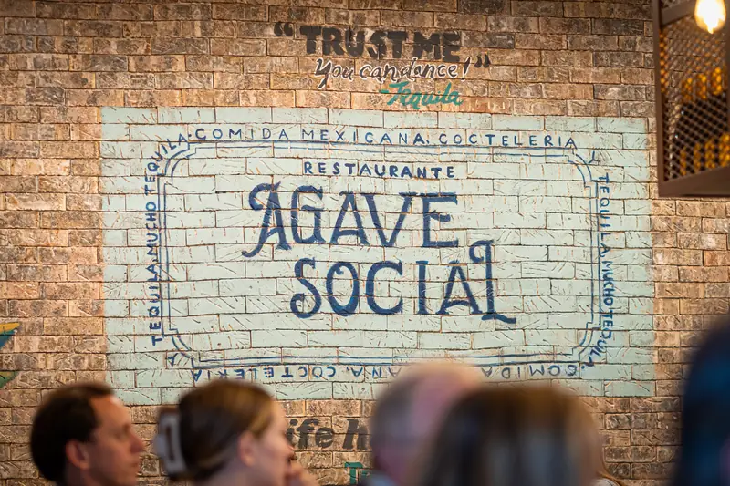 Brick wall mural of the Agave Social badge beneath the painted line Trust me, you can dance, signed Tequila