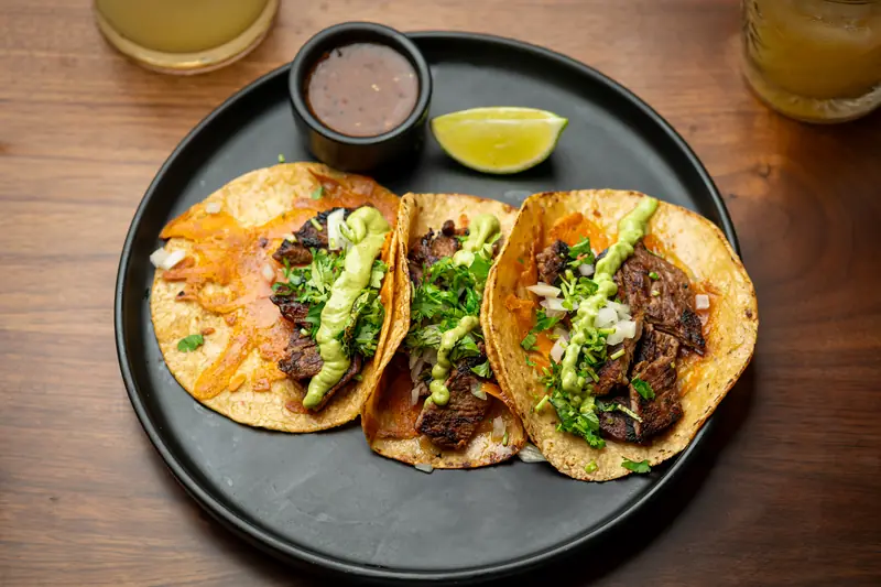 Three birria tacos with shredded beef, avocado crema and cilantro on a black plate with consommé and lime