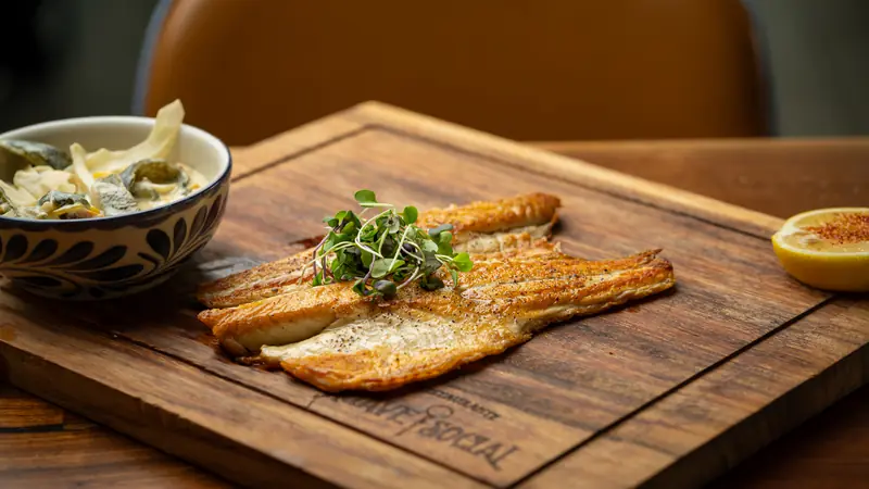 Oven-roasted branzino fillets topped with microgreens on a wooden board with a bowl of rajas poblanas