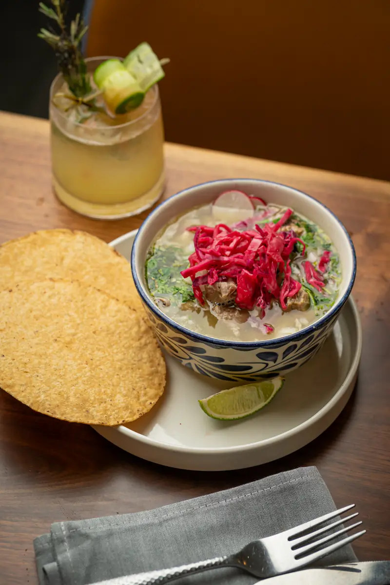 A bowl of pozole topped with pickled red onion and radish, with a margarita and tostadas beside it