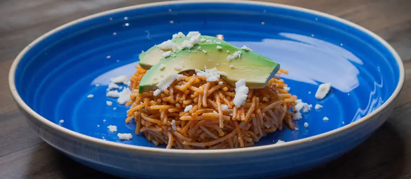 Fideo seco topped with sliced avocado and crumbled queso fresco in a blue bowl