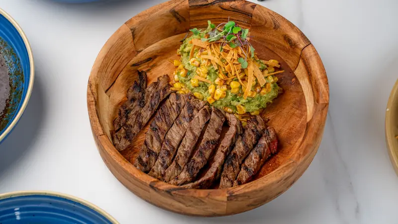 Sliced carne asada with guacamole, corn and shredded cheese in a wooden bowl