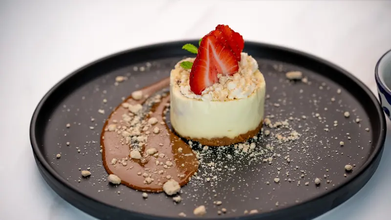 A strawberry-topped cheesecake with caramel and crumble on a dark plate