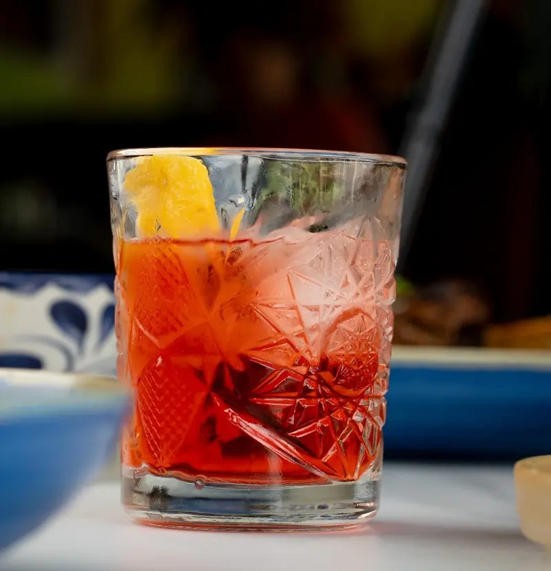 A red cocktail with an orange peel over ice in a cut-crystal glass