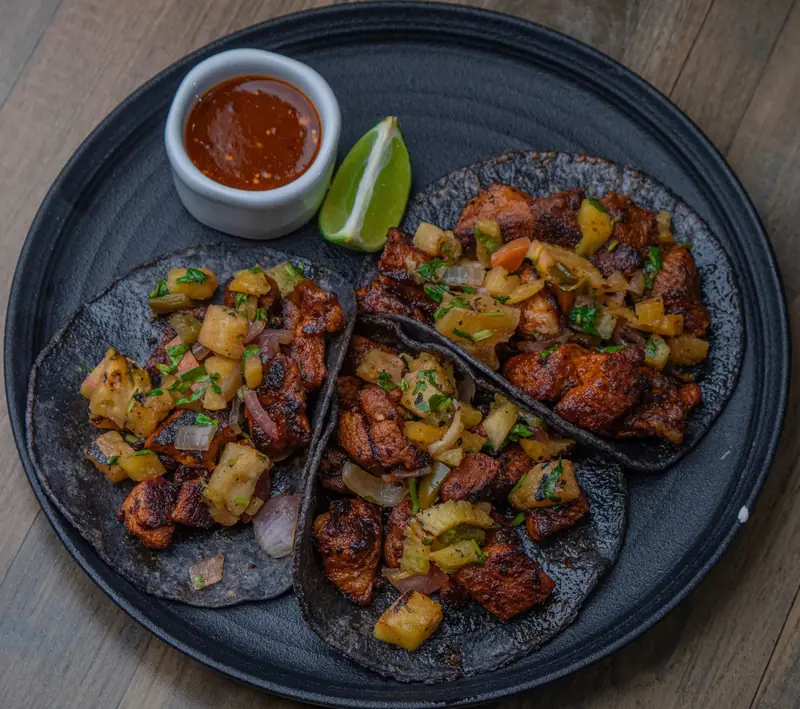 Tacos al pastor with grilled pineapple, lime and salsa on a black plate