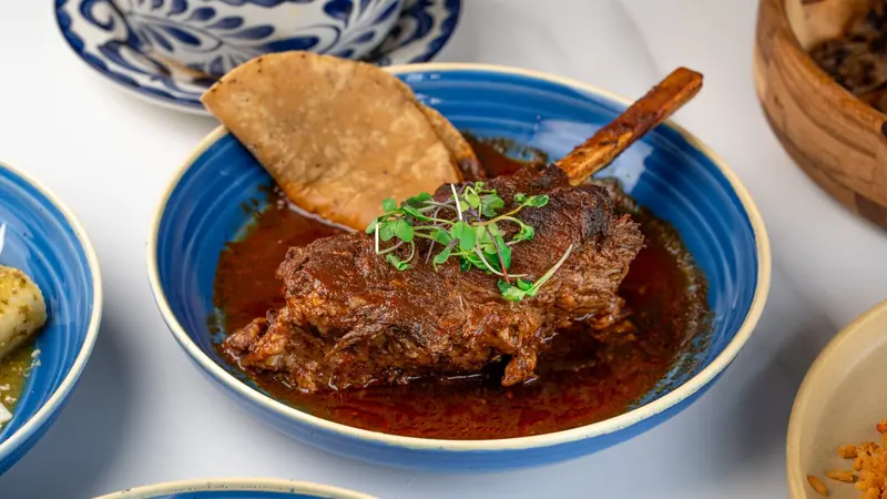 A bone-in braised beef shank in red broth with a tostada, in a blue bowl