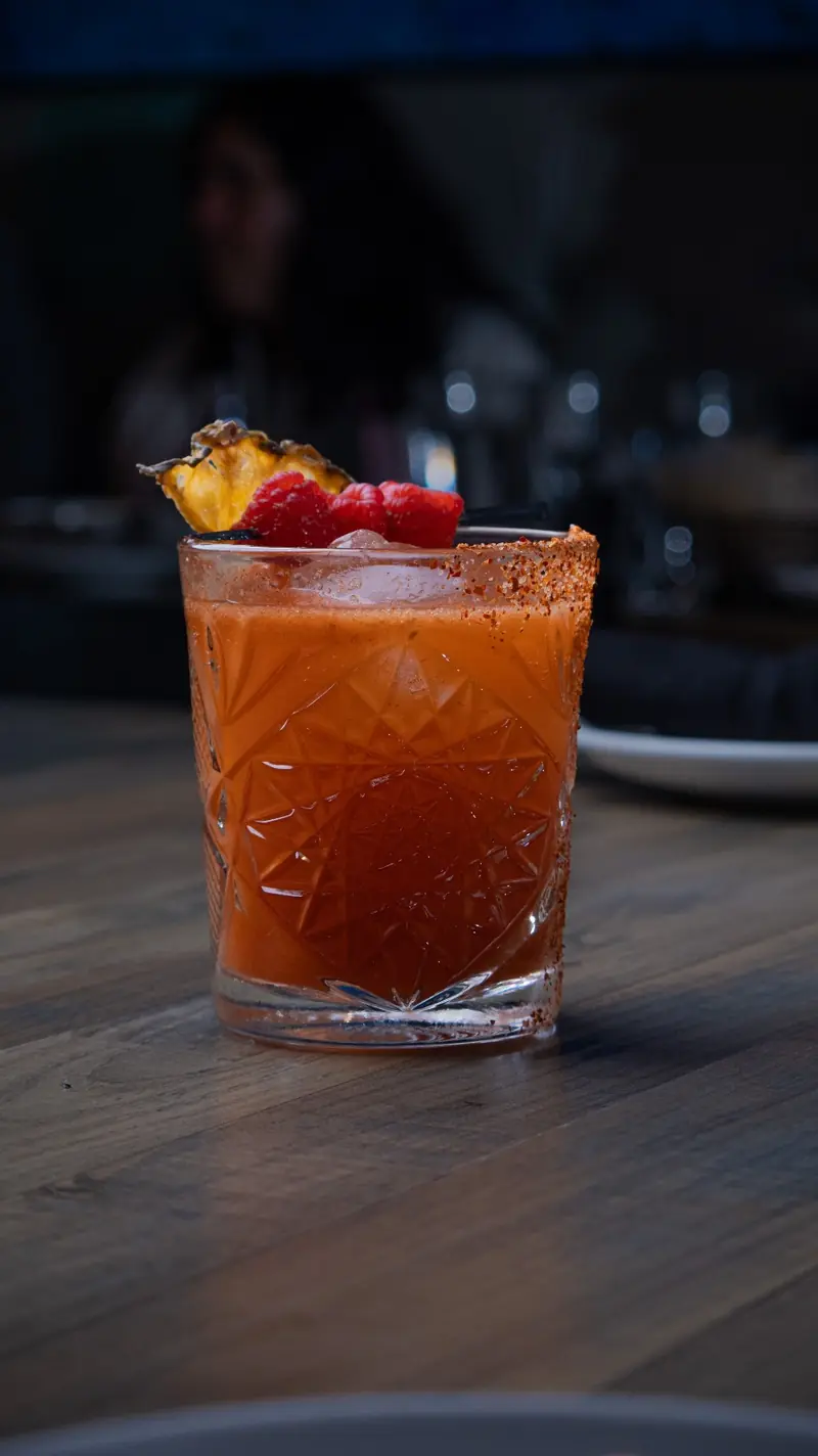A chili-rimmed red cocktail garnished with pineapple and a raspberry
